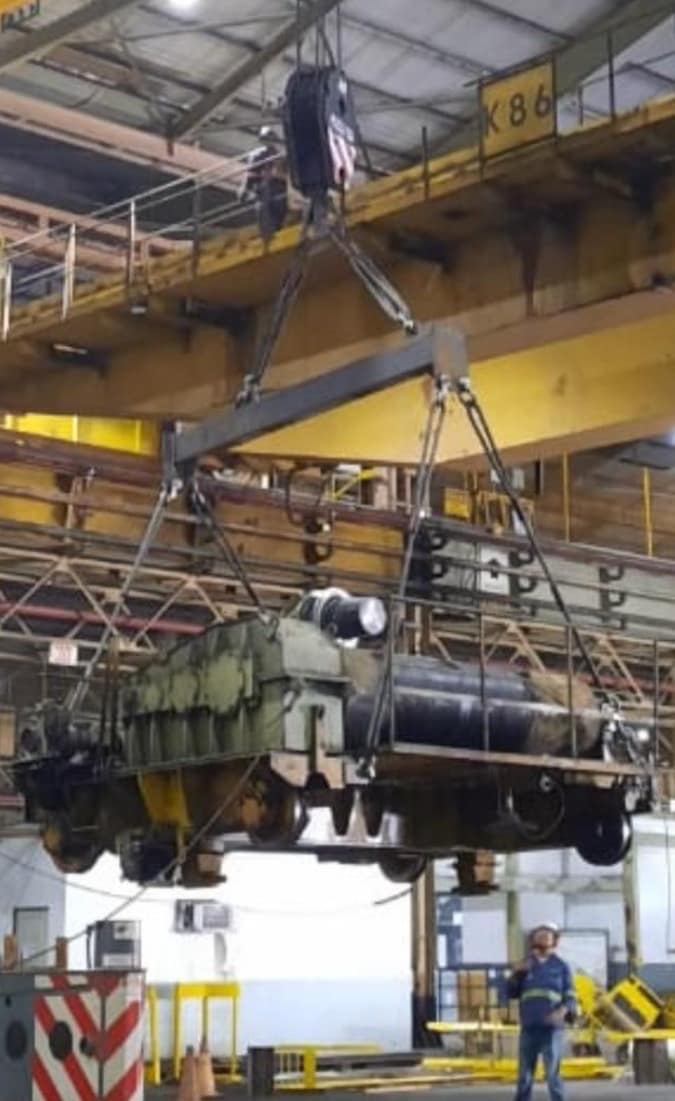 Ponte rolante içando equipamento industrial em galpão fabril, com responsável técnico acompanhando a operação no solo